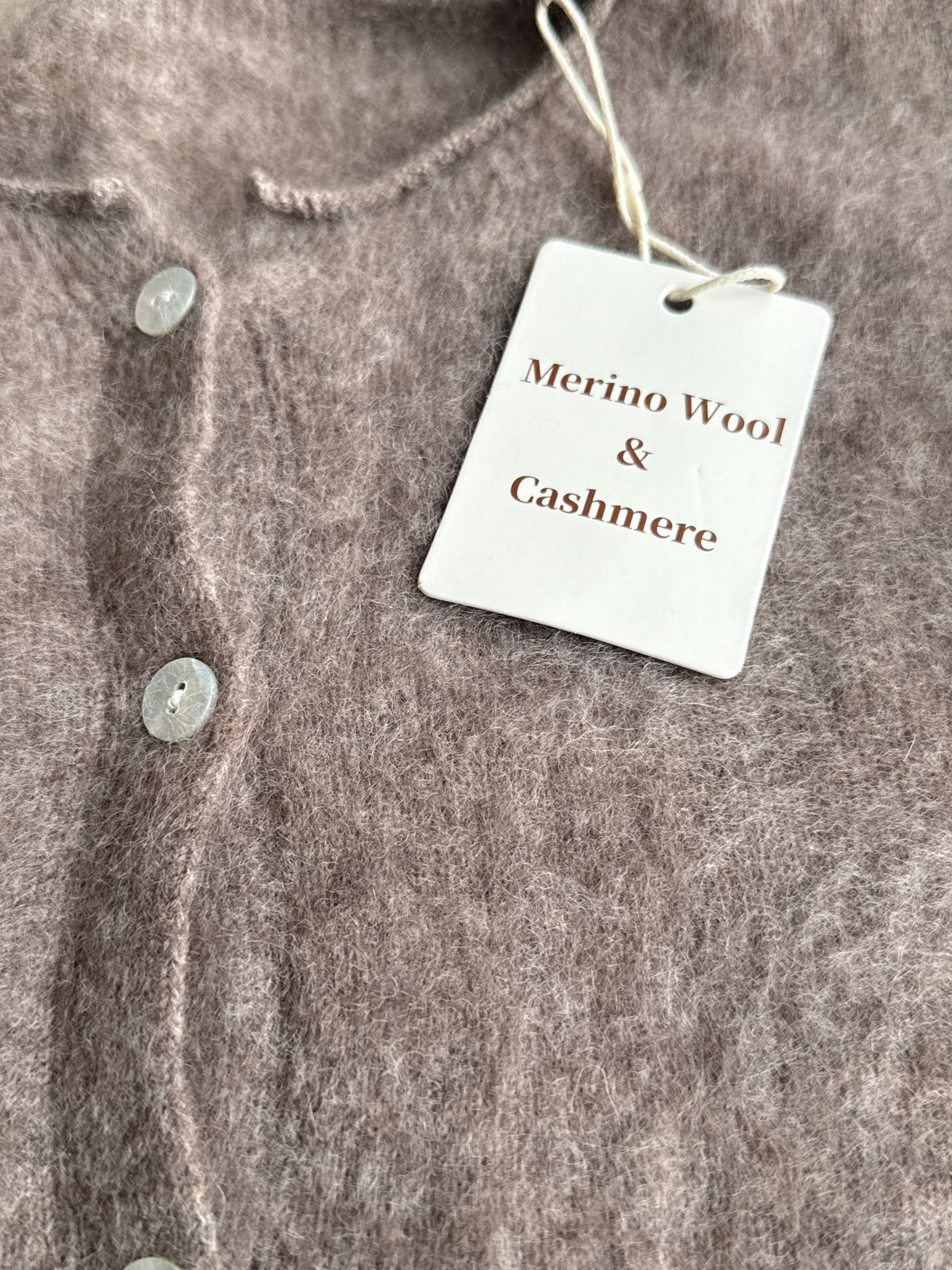 Close-up of a soft, fuzzy knit cardigan in a taupe-brown color, featuring a white paper tag that reads 'Merino Wool & Cashmere'.