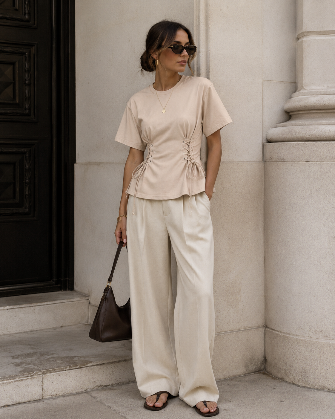 Woman in beige top and white pants standing against a stone wall.