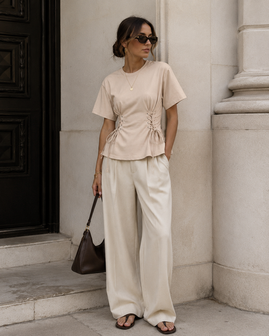 Woman in beige top and white pants standing against a stone wall.