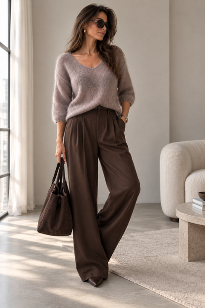 A woman stands in a bright, modern room wearing a fuzzy taupe V-neck sweater, dark brown wide-leg trousers, and holding a dark brown tote bag.