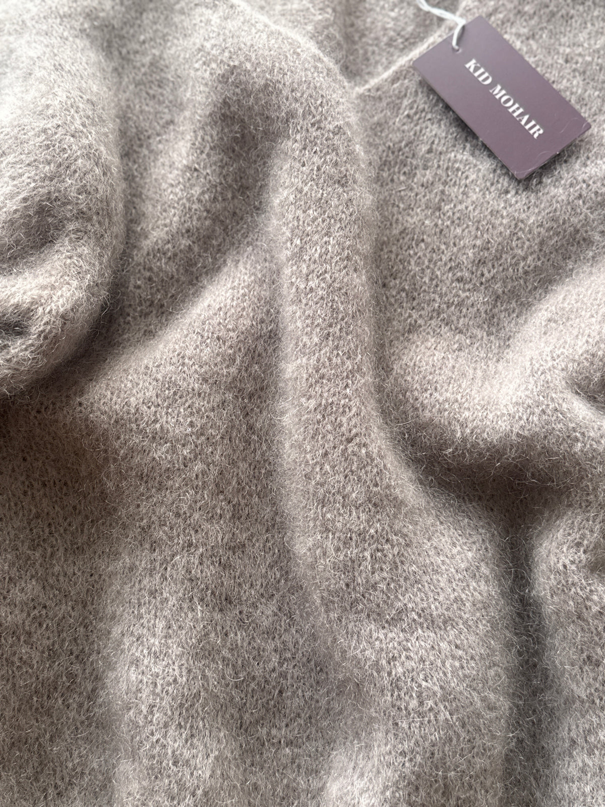 Close-up of a soft, fuzzy knit sweater in a light taupe-gray color, with a dark mauve brand tag reading 'KID MOHLIR' attached near the top right.