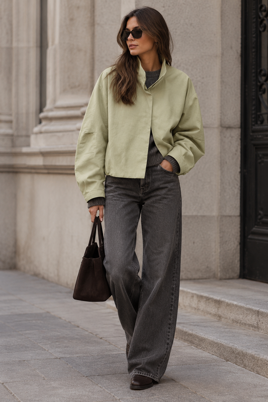 A woman walking outdoors on a stone sidewalk wearing a light sage green cropped jacket, dark gray wide-leg jeans, sunglasses, and carrying a dark brown handbag.