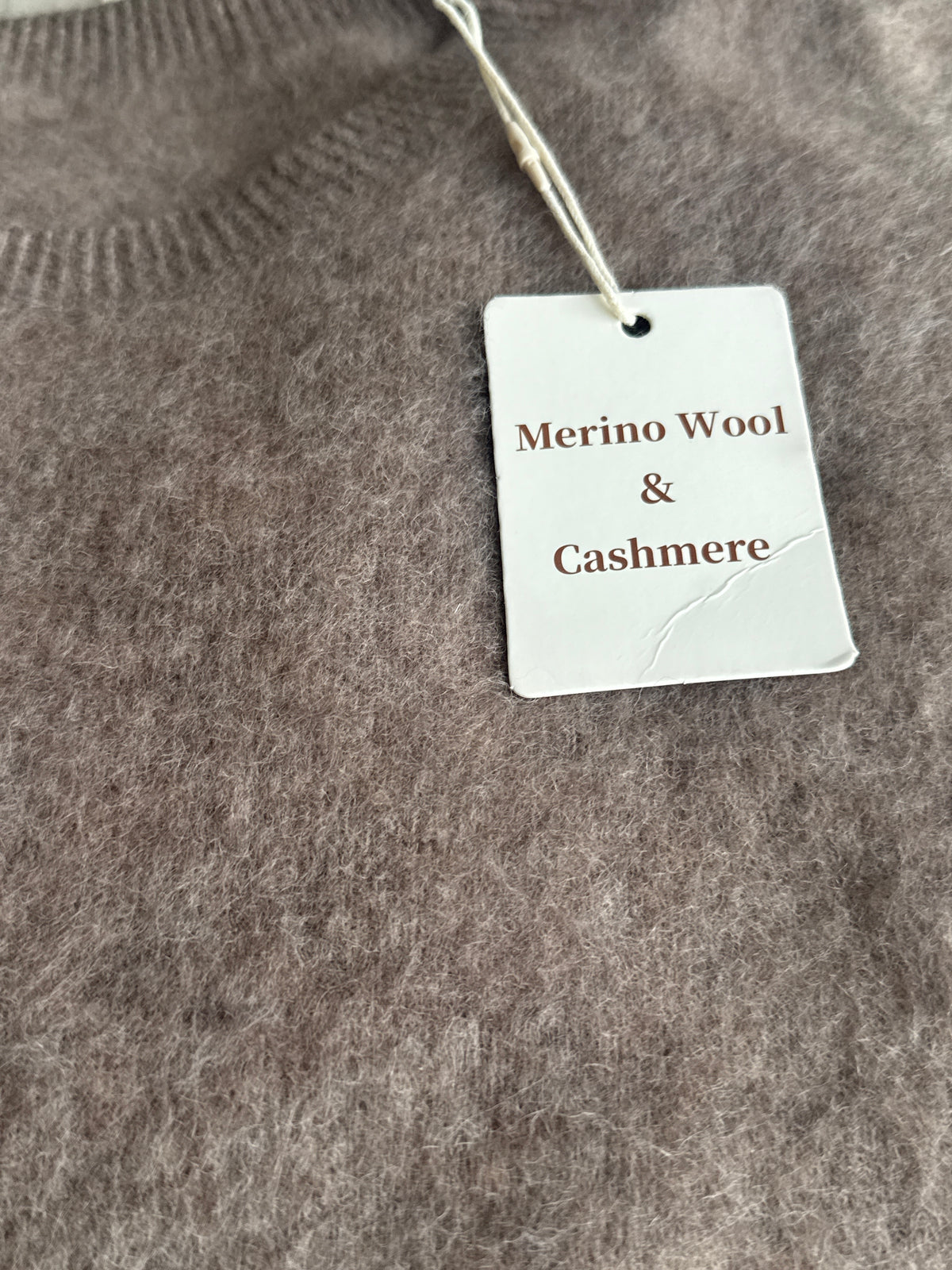 Close-up of a soft, fuzzy knit garment in taupe-brown with a white paper tag reading 'Merino Wool & Cashmere' attached near the neckline.