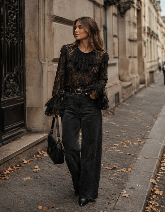 A woman walking on a city sidewalk wearing a black lace long-sleeve blouse with ruffles, black wide-leg jeans, and black pointed shoes, while carrying a black handbag.
