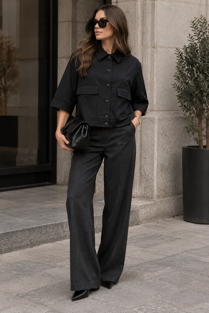 A woman wearing a black cropped button-front shirt with three-quarter sleeves and matching black wide-leg trousers, standing outdoors on a stone walkway and holding a small black clutch.