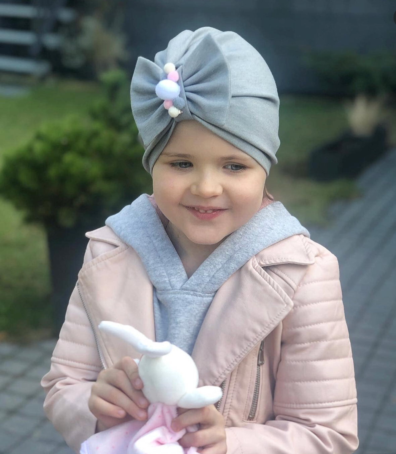 Child wearing a gray headscarf with a bow and holding a stuffed toy outdoors.