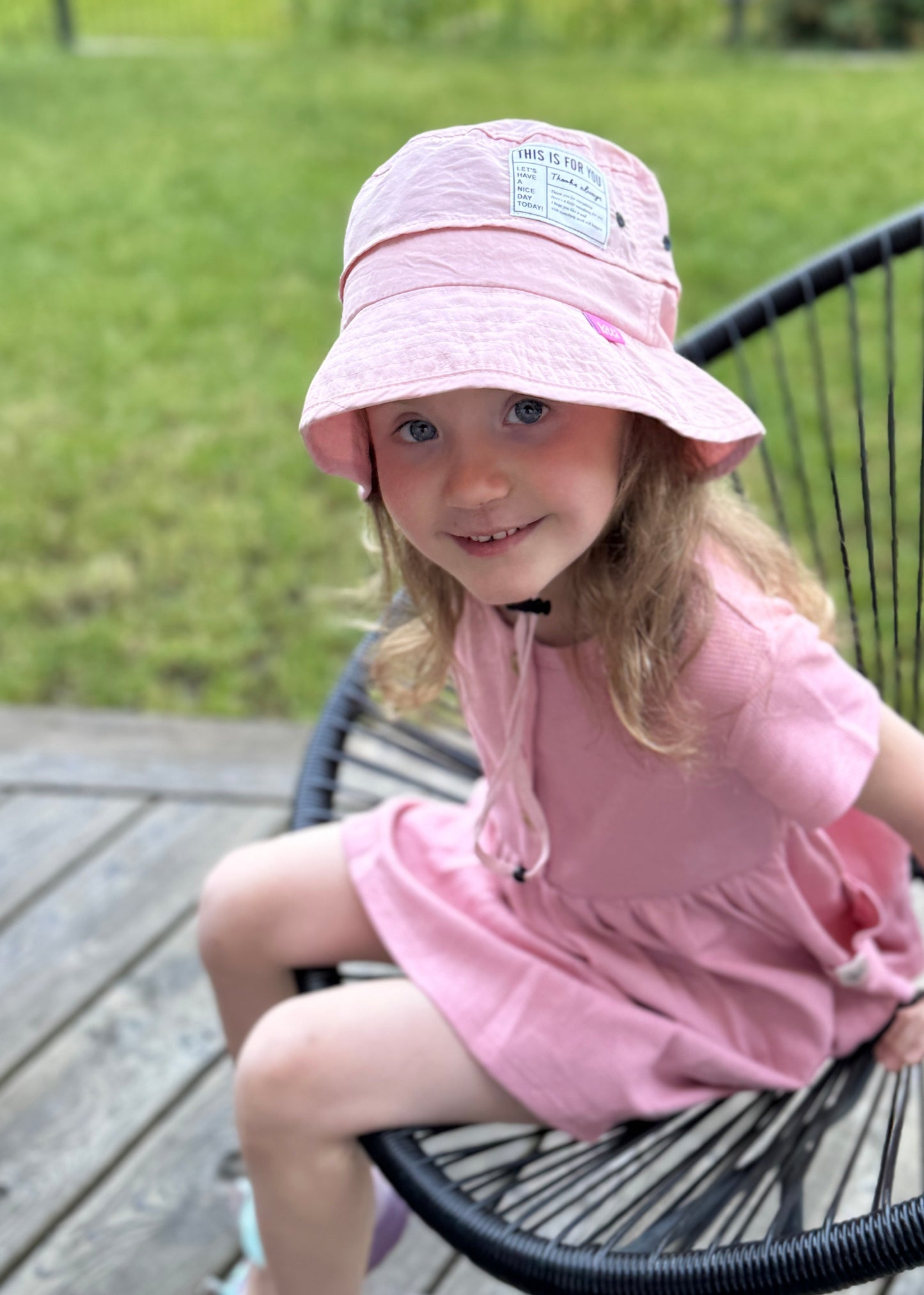 Young girl in a pink dress and hat sitting on a chair outdoors.