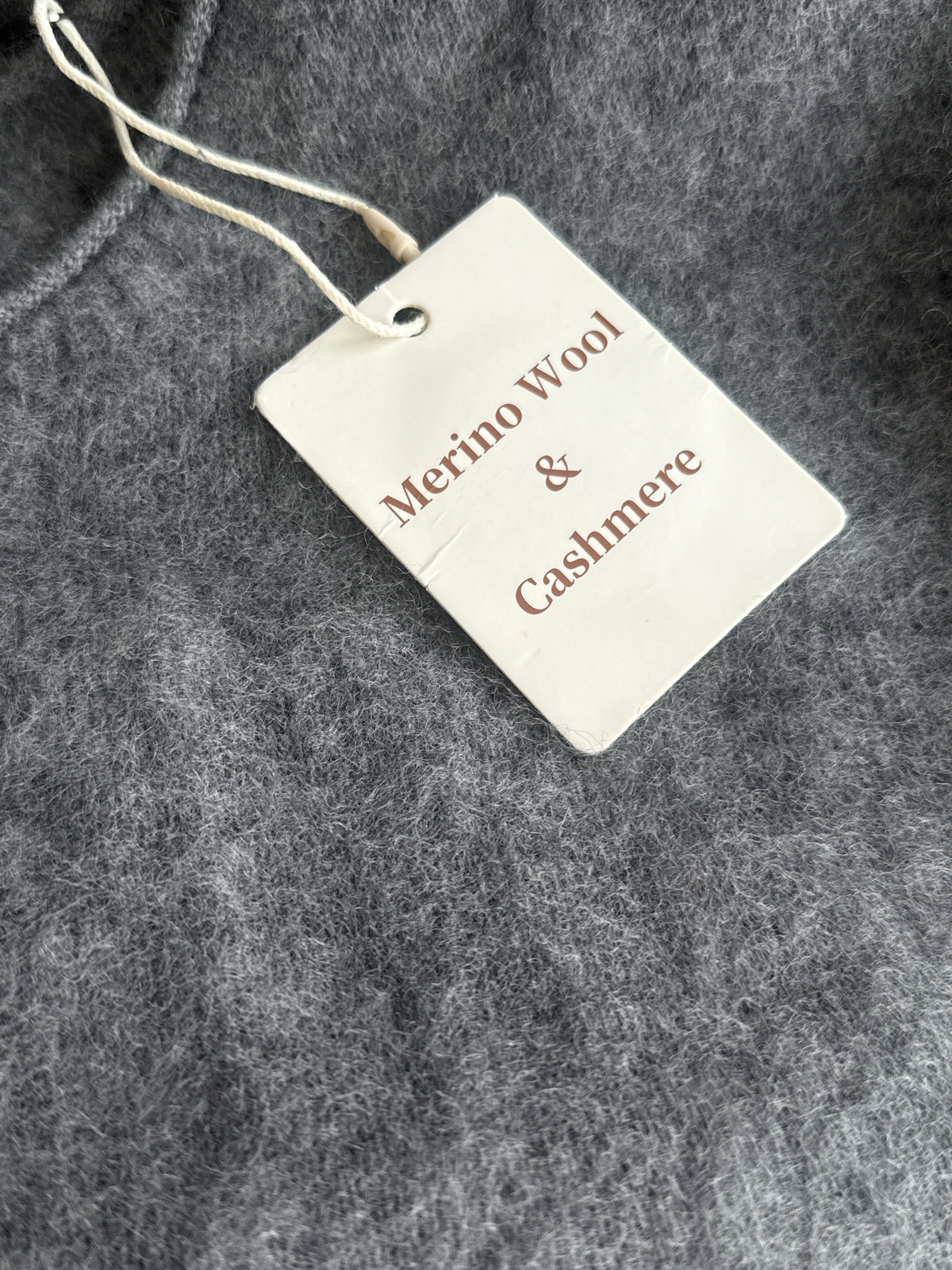 A close-up of a gray textured fabric with a white paper hangtag attached by a white string. The tag reads 'Merino Wool & Cashmere' in brown text.