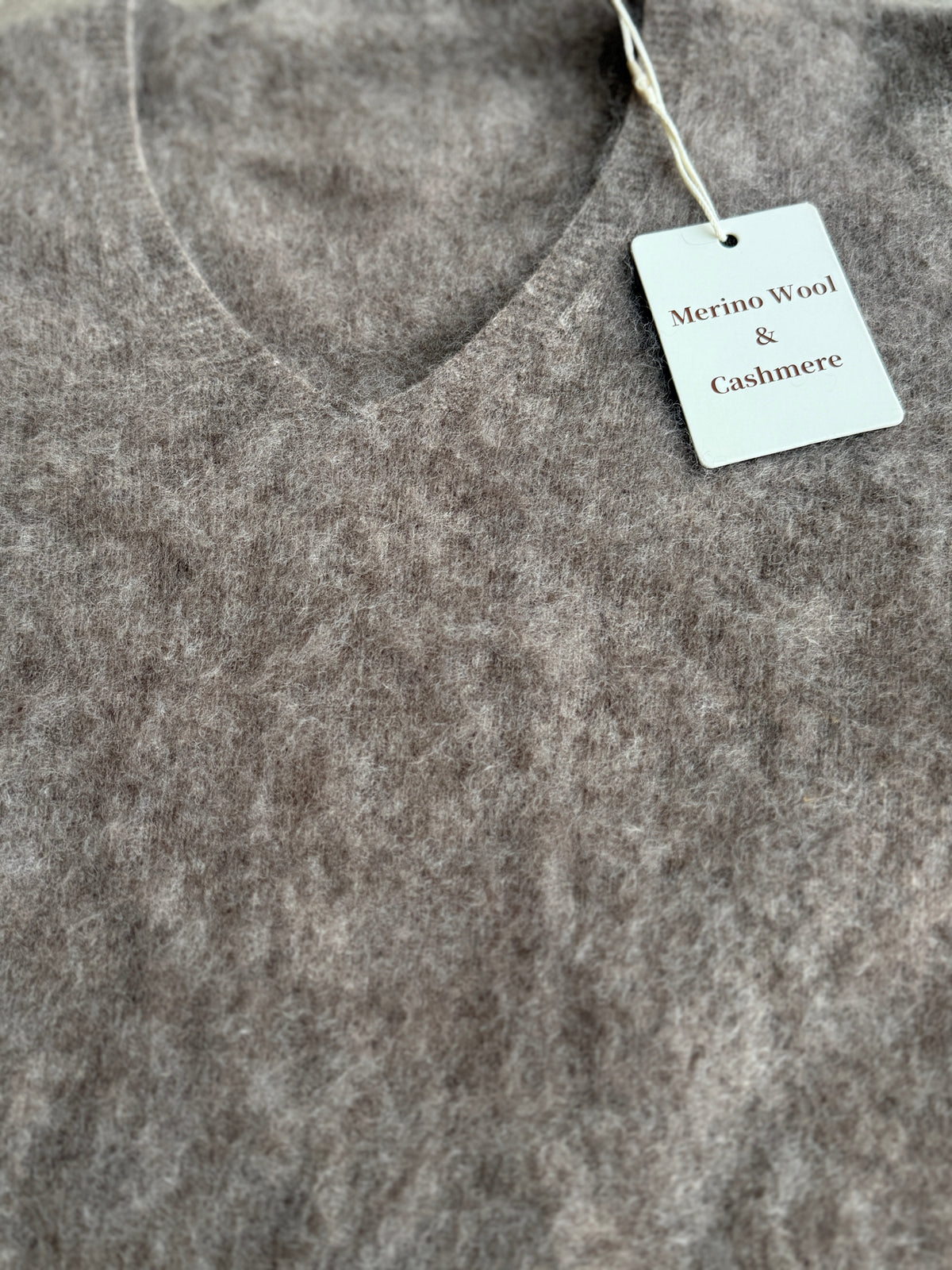 Close-up of a soft, textured grayish-brown knit garment with a white paper tag reading 'Merino Wool & Cashmere' attached near the neckline.