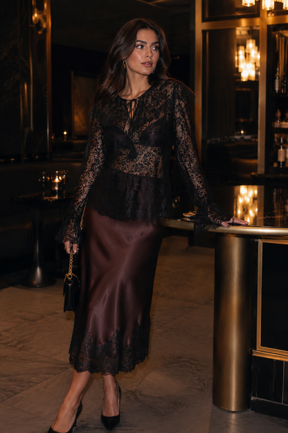 Woman in a dark lace top and satin skirt standing in an elegant indoor setting.