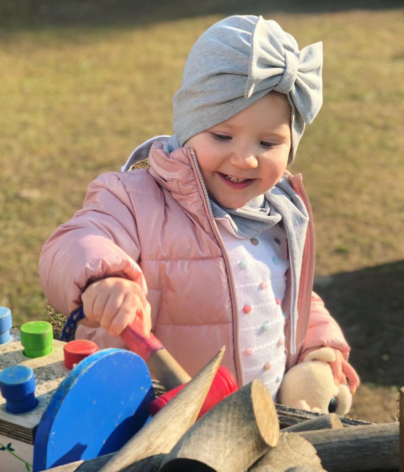 Child playing with toys outdoors wearing a pink jacket and gray headband with a bow.