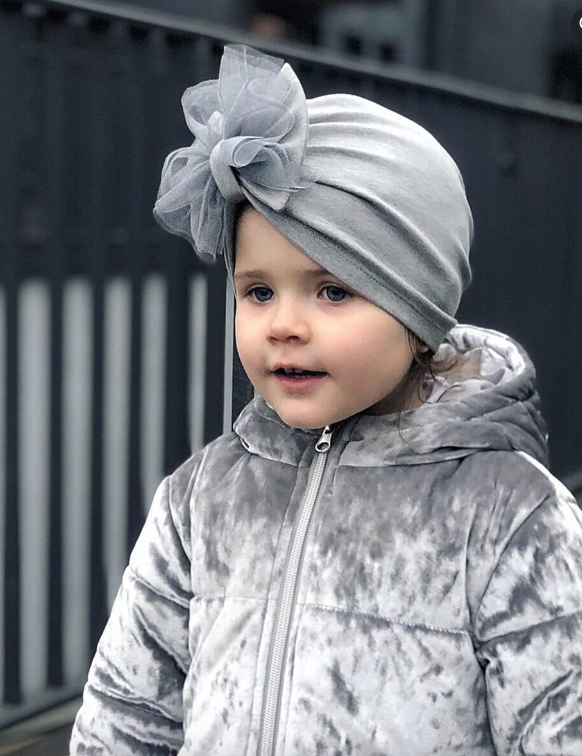 Child wearing a gray turban with a large flower accessory outdoors.