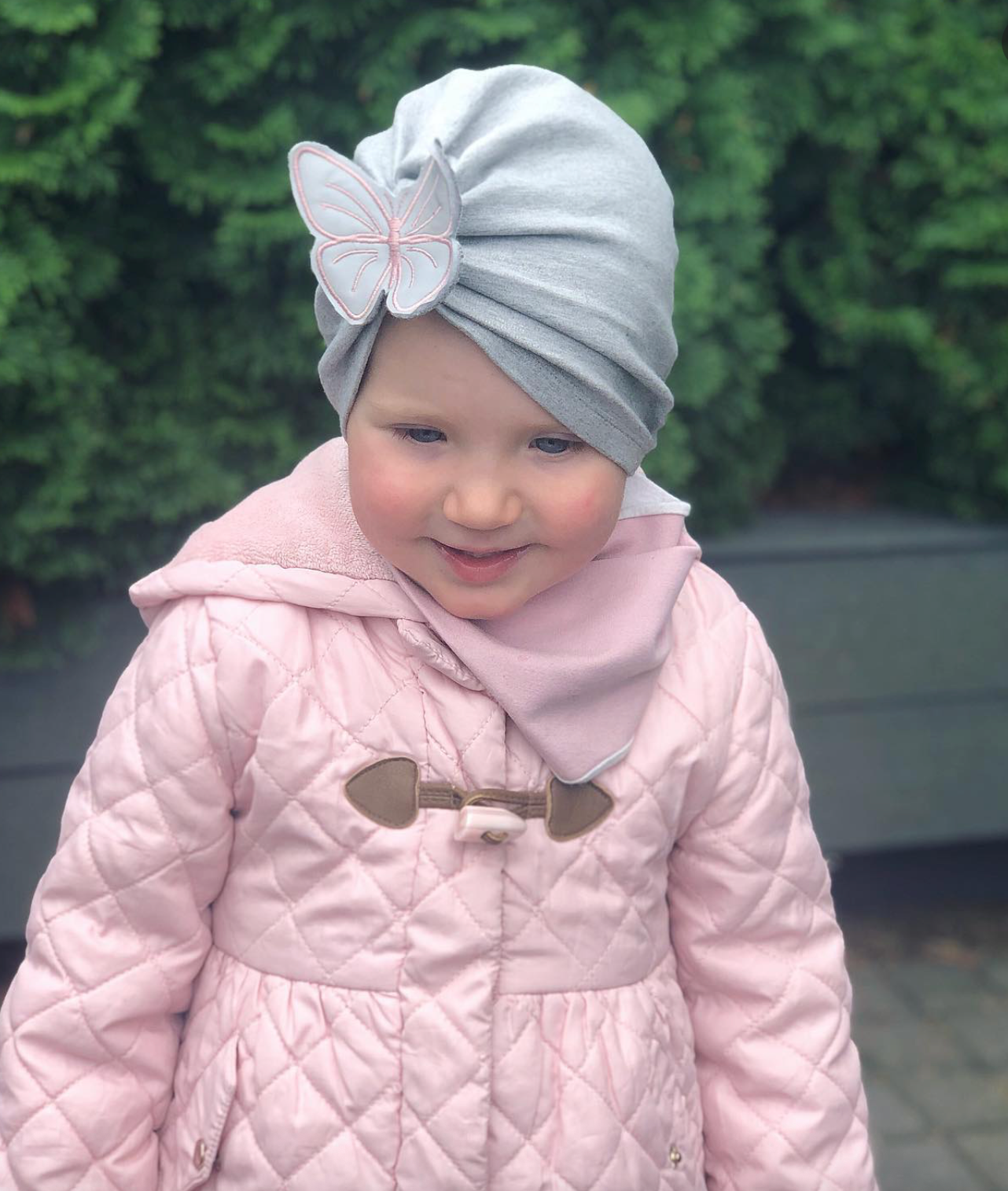 Child wearing a light gray turban with a butterfly design and a pink quilted coat.