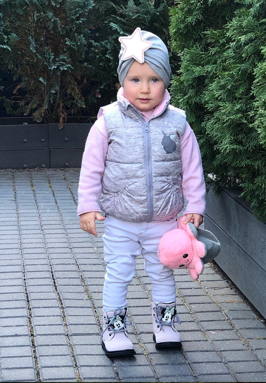 Child in winter clothing with a pink toy, standing on a paved area with greenery in the background.