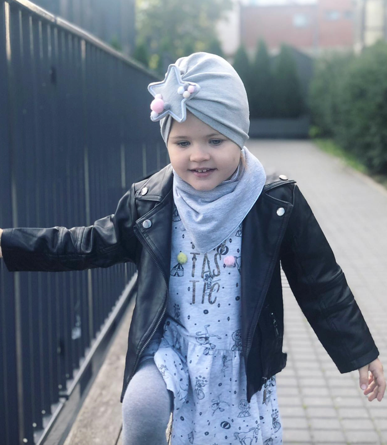 Child wearing a gray headscarf and black leather jacket outdoors.